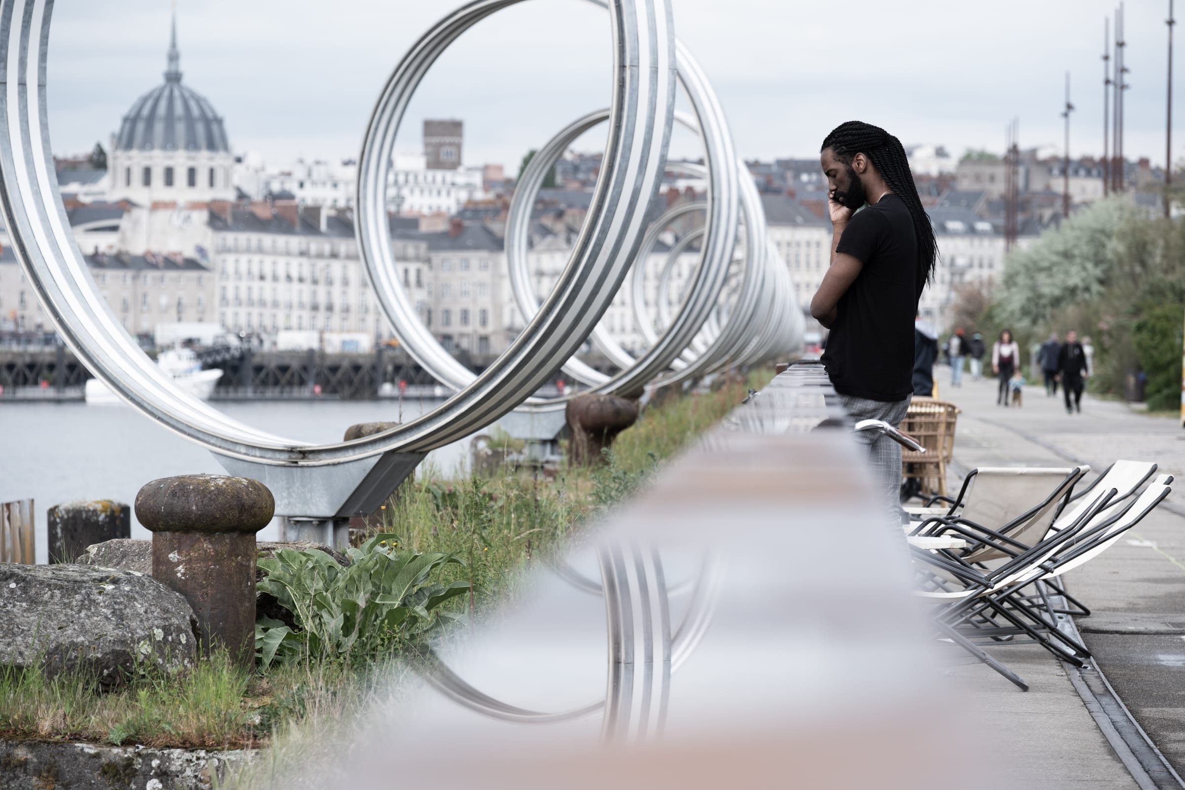 Homme au téléphone à travers une sculpture en anneaux d'acier sur les quais de Nantes, dôme du Bouffay en arrière-plan